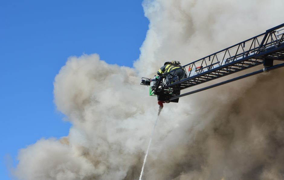 Firefighter in full turnout gear on an aerial ladder directing a stream of water into heavy smoke