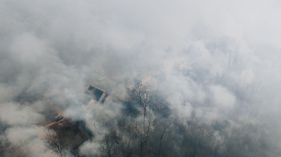 Aerial photograph of a residential neighborhood surrounded by dense forest with smoke from wildfire visible across the landscape