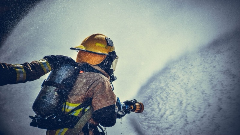 Firefighter in protective gear spraying a wide fan of water during a live fire operation