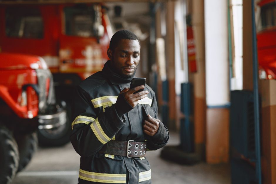 Firefighter in uniform checking his smartphone inside a fire station with fire trucks in the background