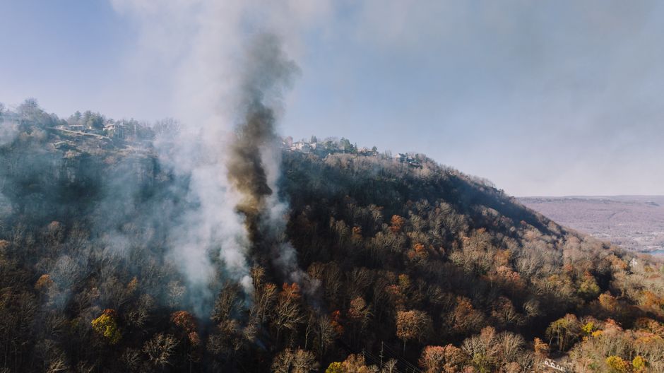 Aerial shot of smoke rising from a forest fire on a hillside near homes